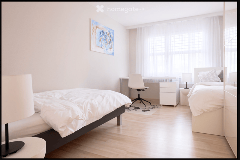 Modern bedroom with two beds, nightstands, a desk with a chair, and a large mirror. Light-colored bedding and furniture, with a painting on the wall and large windows with sheer curtains.