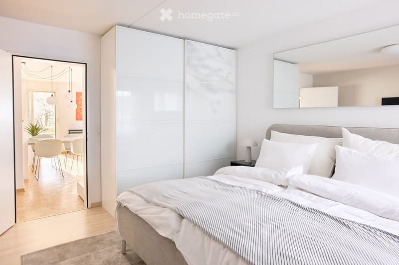 Modern bedroom with a large bed, nightstand, lamp, and sliding wardrobe. The room features light-colored walls, wooden flooring, and a view into an adjacent dining area with chairs and a table.