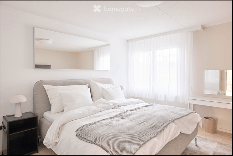 Modern, minimalist bedroom with a large bed, white bedding, a nightstand with a lamp, and a large mirror on the wall.
