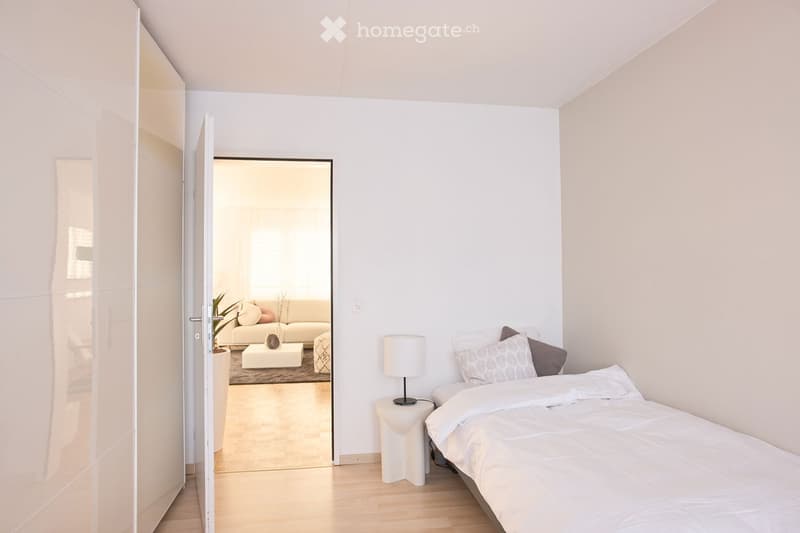 Modern bedroom with a large bed, nightstand, lamp, and an open door leading to a living area with a sofa and coffee table.