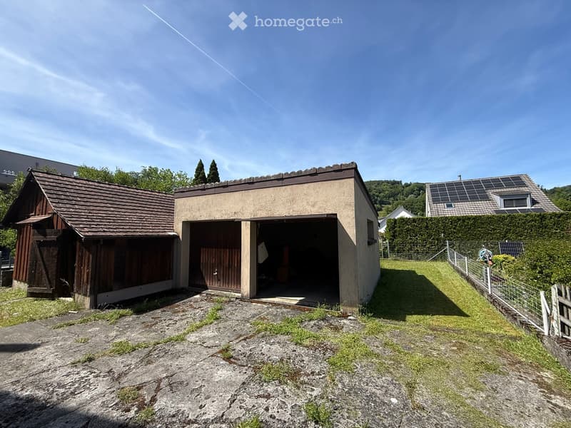 A small, single-story building with a garage and a wooden shed attached, surrounded by a grassy area and a fence.