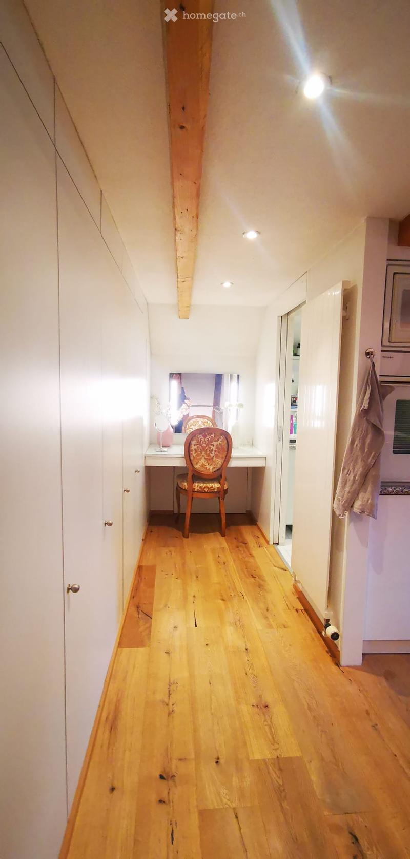 A narrow hallway with wooden flooring, white walls, and built-in cabinets on one side. A small vanity area with a chair and mirror is visible at the end of the hallway.