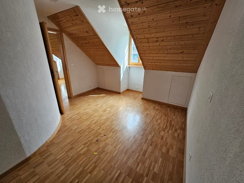 Empty attic room with wooden flooring, slanted wooden ceiling, and large windows allowing natural light. The space is unfurnished with visible electrical outlets and a small built-in cabinet.