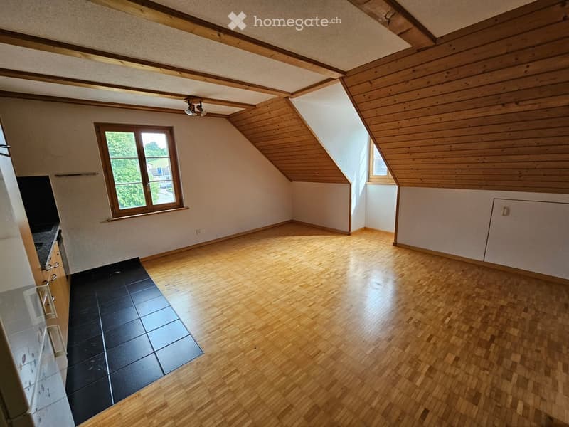 Empty attic room with wooden floors, exposed wooden beams, and large windows allowing natural light. The space is unfurnished and features a mix of wooden and tiled flooring.