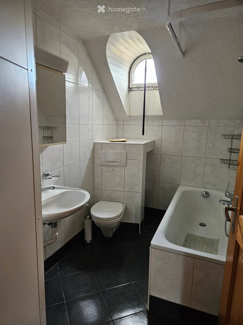Modern bathroom with a white sink, toilet, bathtub, and a mirror. The room features dark tiled flooring and a skylight window providing natural light.
