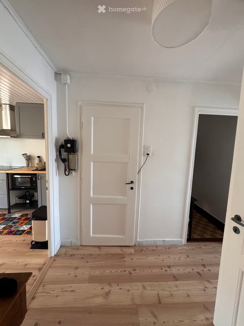 Empty room with wooden flooring, white walls, and a white door. The adjacent kitchen area is visible with cabinets, a microwave, and a countertop.