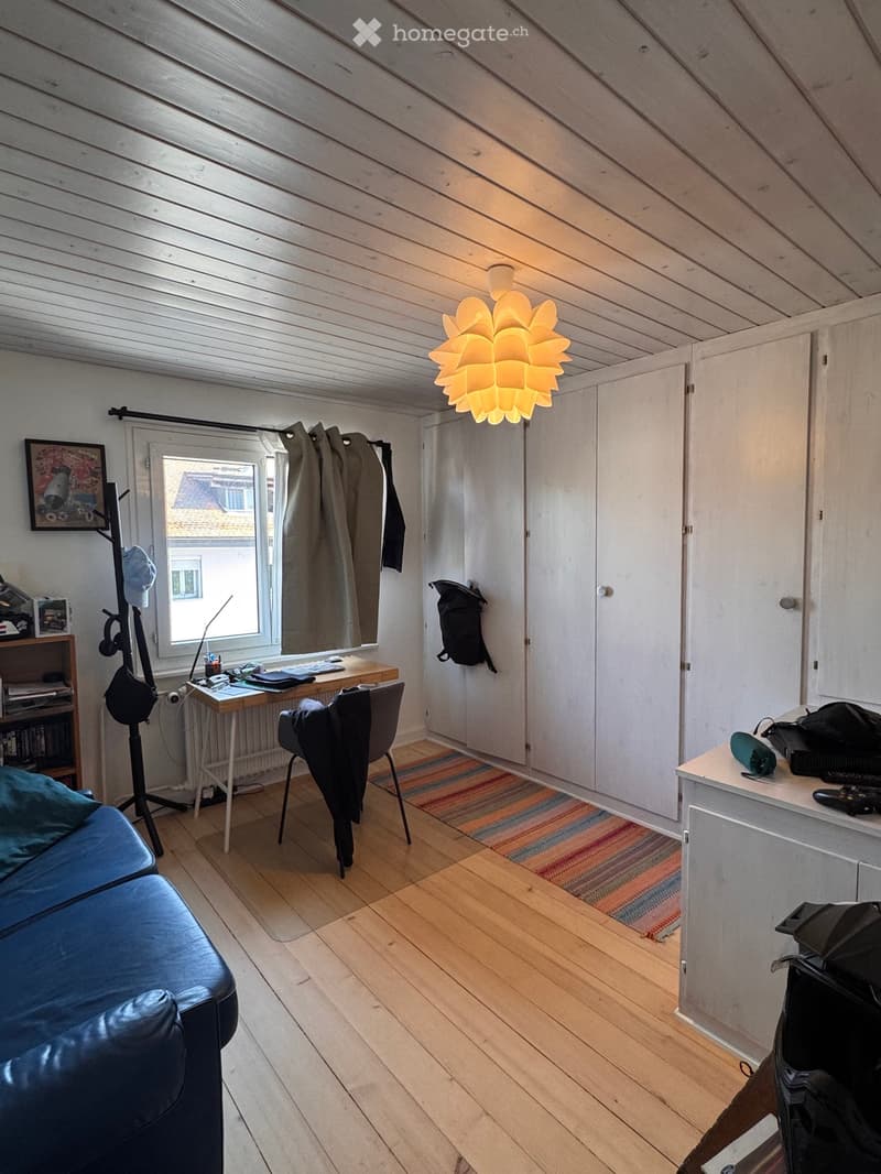Cozy attic room with wooden floors, slanted ceilings, and modern furnishings including a blue leather sofa, a desk with a chair, a colorful rug, and a decorative lamp.