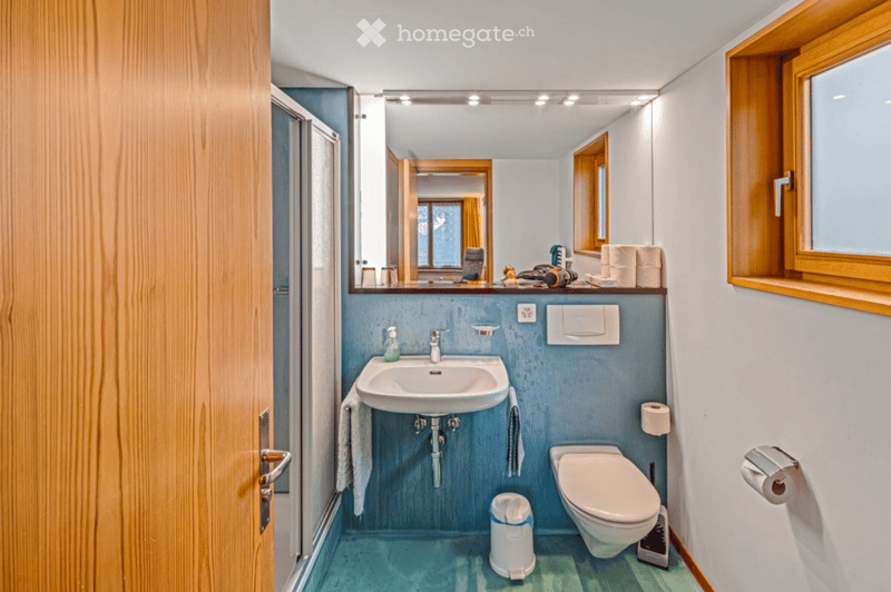 A small, modern bathroom with a white sink, toilet, and mirror. The walls are painted in a light blue color, and the floor is covered with turquoise tiles. There are towels hanging on the sides of the sink, and a trash bin is placed near the toilet.