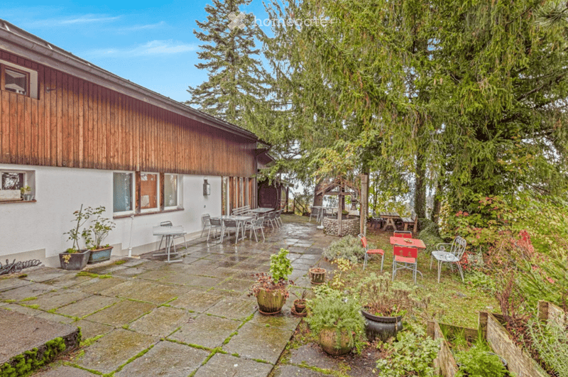 A well-maintained garden area with a stone-paved patio, outdoor furniture, potted plants, and a wooden house with large windows and a wooden facade.