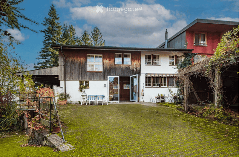 A two-story house with a wooden facade and white walls, featuring large windows and a covered entrance. The surrounding area includes a moss-covered ground, outdoor furniture, and greenery.