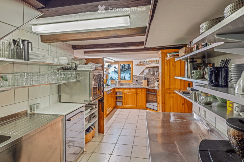 A well-equipped kitchen with wooden cabinets, stainless steel appliances, and shelves filled with dishes and glassware. The space features a tiled floor and a large window offering a view of the outdoors.