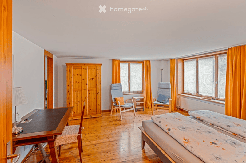 Bright bedroom with wooden flooring, two single beds, a desk with a chair, two armchairs, a wardrobe, and large windows with orange curtains.