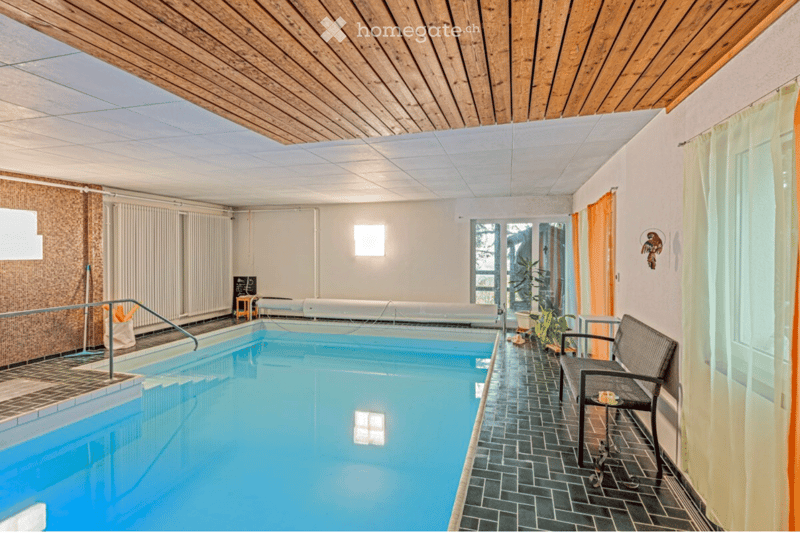 Indoor swimming pool area with wooden ceiling, tiled walls, and a lounge chair near the pool.