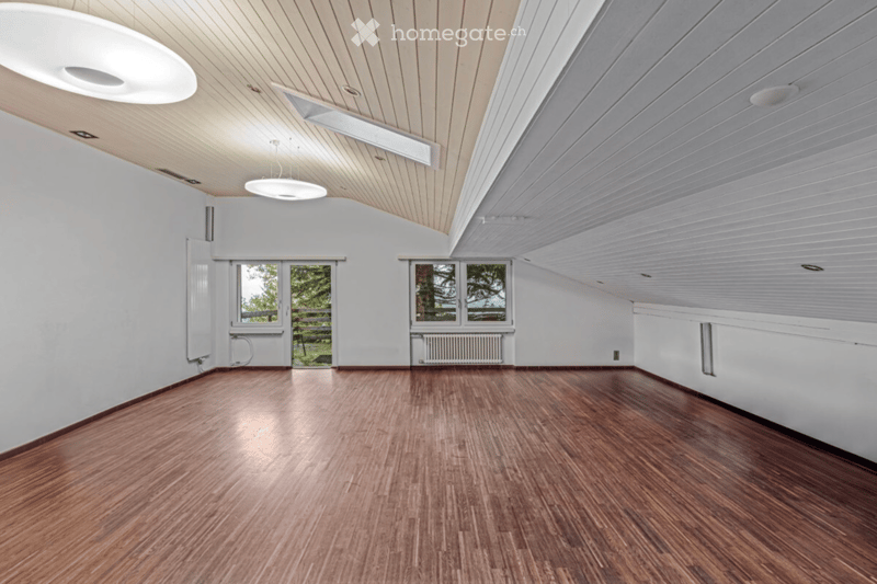 Empty room with wooden flooring, white walls, and large windows offering a view of the outdoors. The ceiling features a slanted design with recessed lighting.