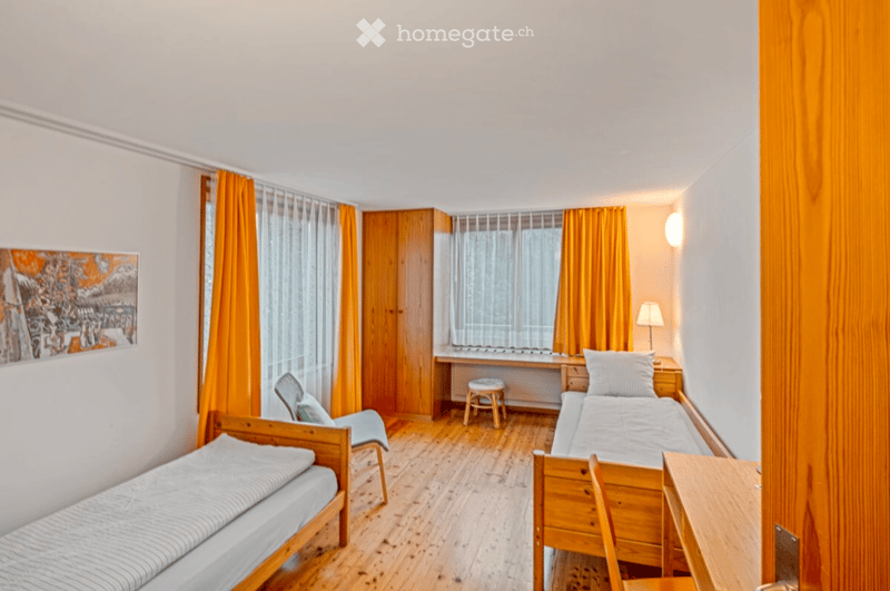 Cozy, well-lit bedroom with wooden floors, two single beds, a small table, a chair, and a stool near the window. Orange curtains and a painting add color to the space.