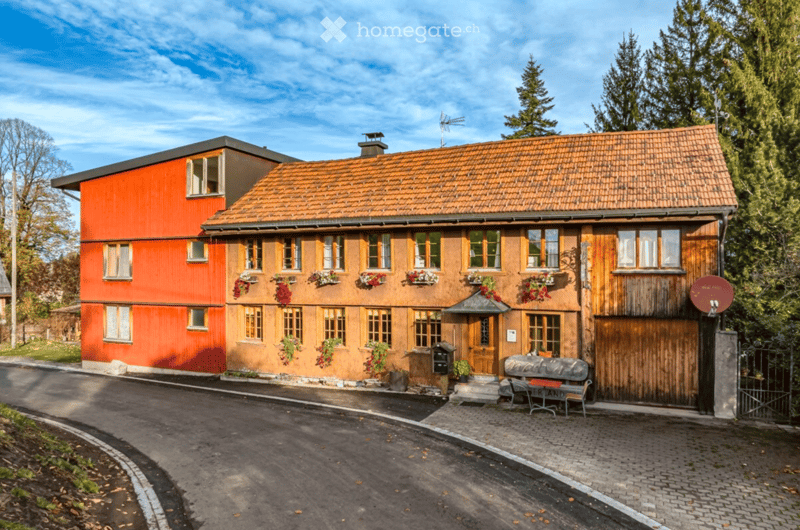 A two-story house with a combination of orange and wooden facades, multiple windows with flower boxes, and a small entrance area with a bench.