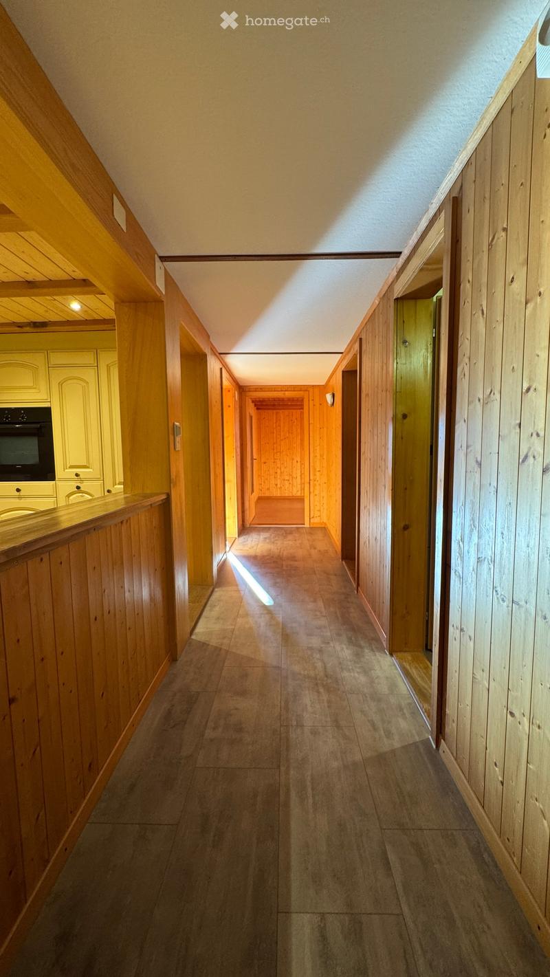 A long, narrow hallway with wooden panel walls and a tiled floor, featuring several doors and a visible kitchen area on the left side.