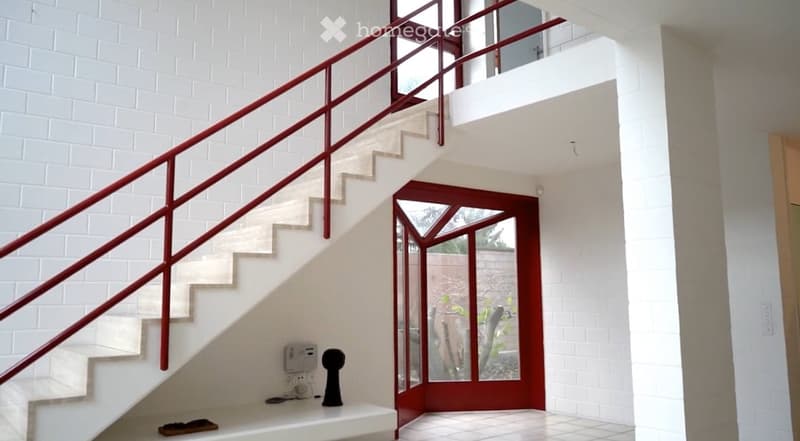 Modern hallway with white brick walls, a staircase with red metal railing, and a glass-paneled door leading outside.