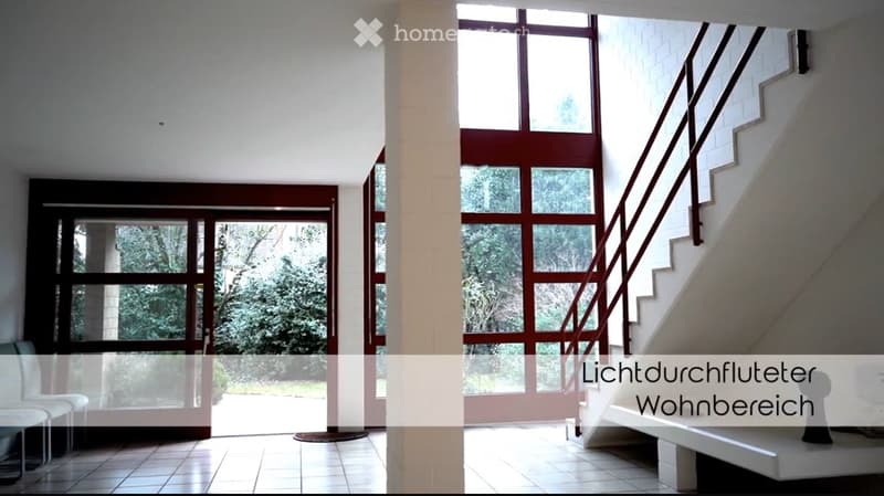 Bright, modern hallway with large windows and a staircase leading upwards. The space features white walls, tiled flooring, and a central column.