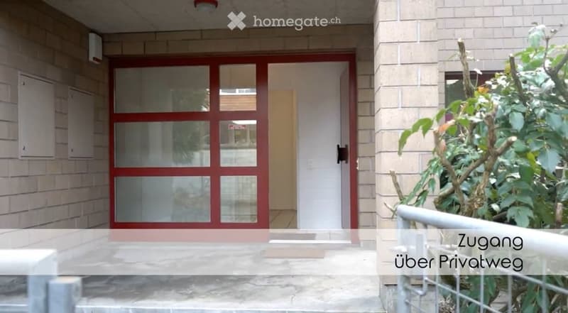 Entrance to a building with a red-framed glass door, white walls, and a small garden area with plants and a metal railing.