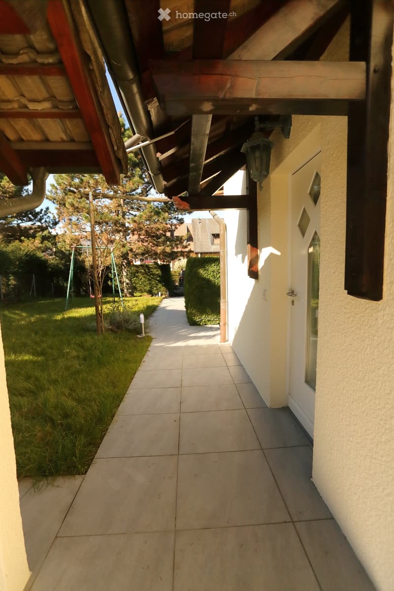 Corridor / Hallway. A covered walkway leading to a house entrance, with a swing set and greenery in the background.