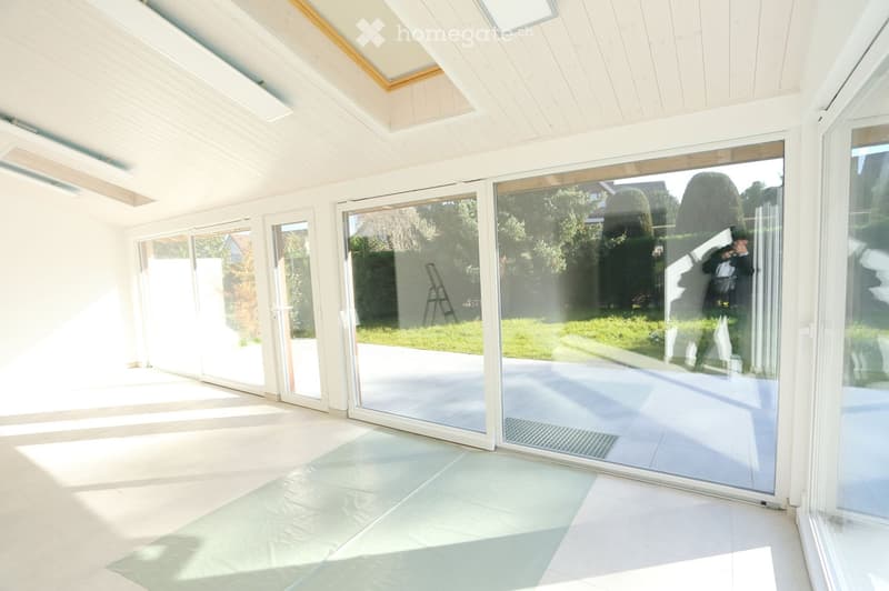 Unfurnished Room. An empty room with white walls, ceiling, and floor. It has large glass doors and windows, and a plastic sheet on the floor. There is a reflection of a person and a ladder outside.