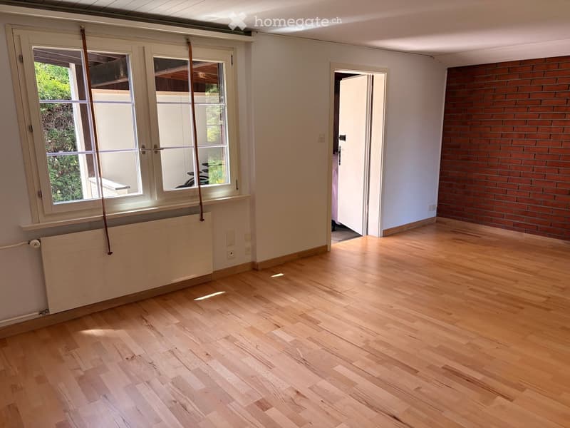 Empty room with wooden flooring, white walls, large windows, and a radiator. There is an open doorway leading to another room with a brick wall visible.