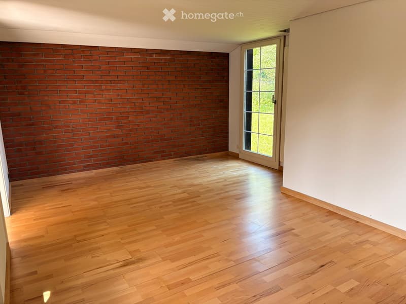Empty room with wooden flooring and a brick accent wall, featuring a large window with a view of greenery outside.