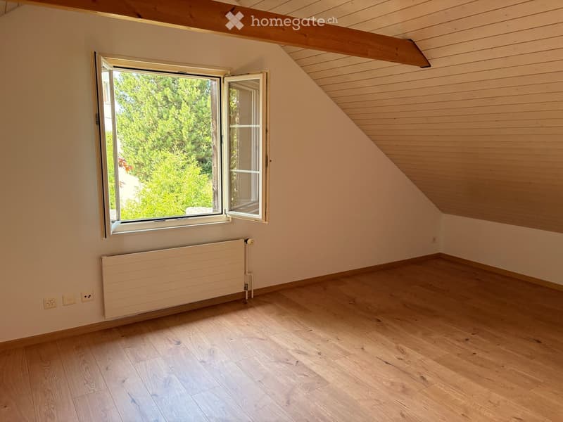 Empty attic room with wooden flooring, exposed wooden beams, large window with view of greenery, radiator, and electrical outlets.