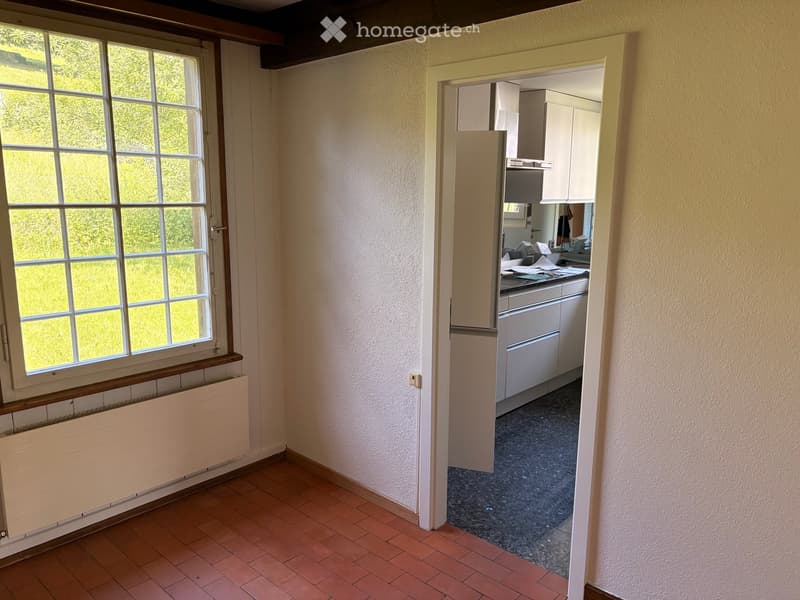 Empty room with tiled floor, large window with white frame, radiator below window, and an open doorway leading to a kitchen area with white cabinets and appliances.