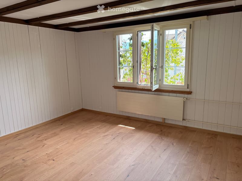 Empty room with light wooden walls, wooden floor, and three windows with white frames. A radiator is mounted below the windows.