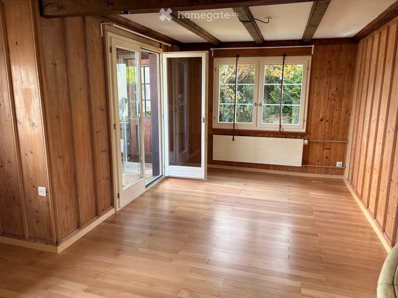 Empty room with wooden walls and floor, large windows, and sliding glass doors leading outside.