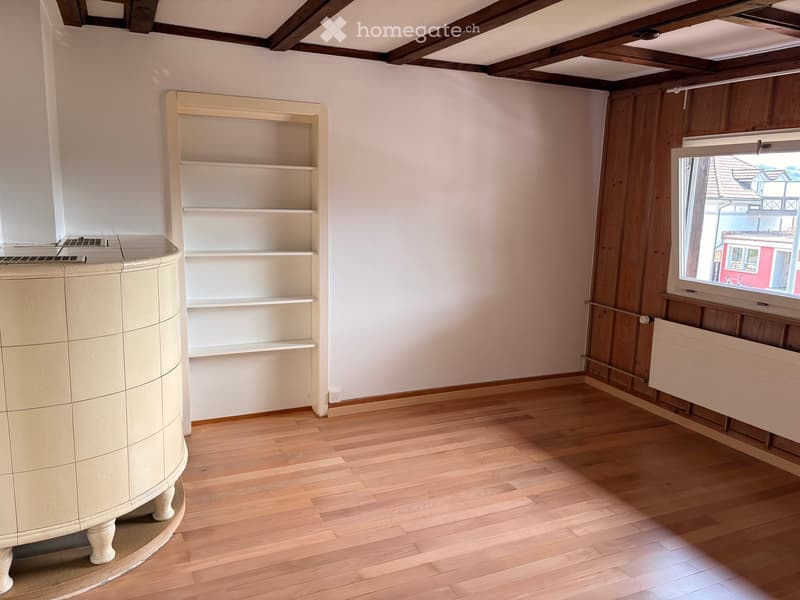 Empty room with wooden flooring, white walls, exposed wooden beams on the ceiling, a built-in shelf unit, and a window showing a view of the street.