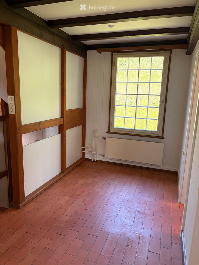 Empty room with exposed wooden beams, red brick floor, white walls, radiator, and a window with multiple panes.