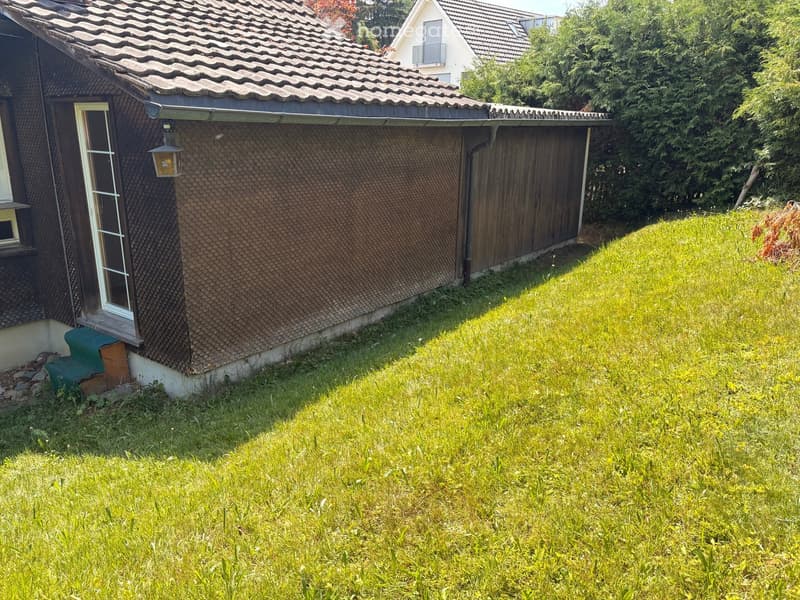 A small, single-story house with a brown tiled roof and textured exterior wall, surrounded by a grassy yard and trees.