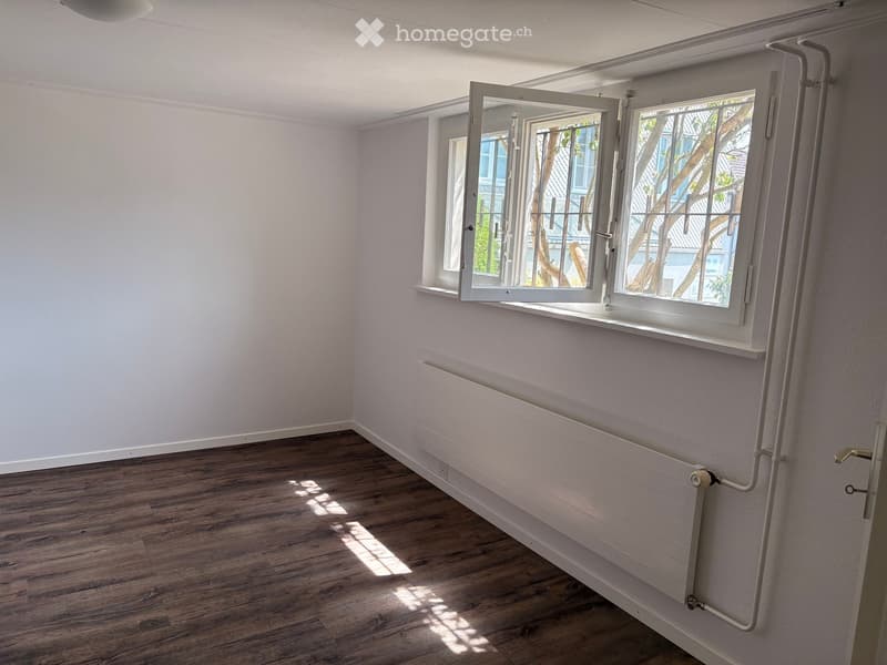 Empty room with wooden flooring, white walls, and a large window with white frame and radiator below it.
