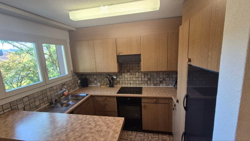 The kitchen features light wooden cabinets, a tiled backsplash, and a countertop with a sink, faucet, and small appliances. There is also an oven and refrigerator visible.