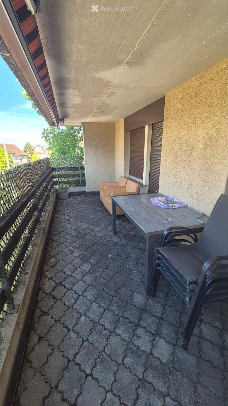 Balcony with a table, chairs, and a small sofa, overlooking a fenced yard with trees and a house in the background.