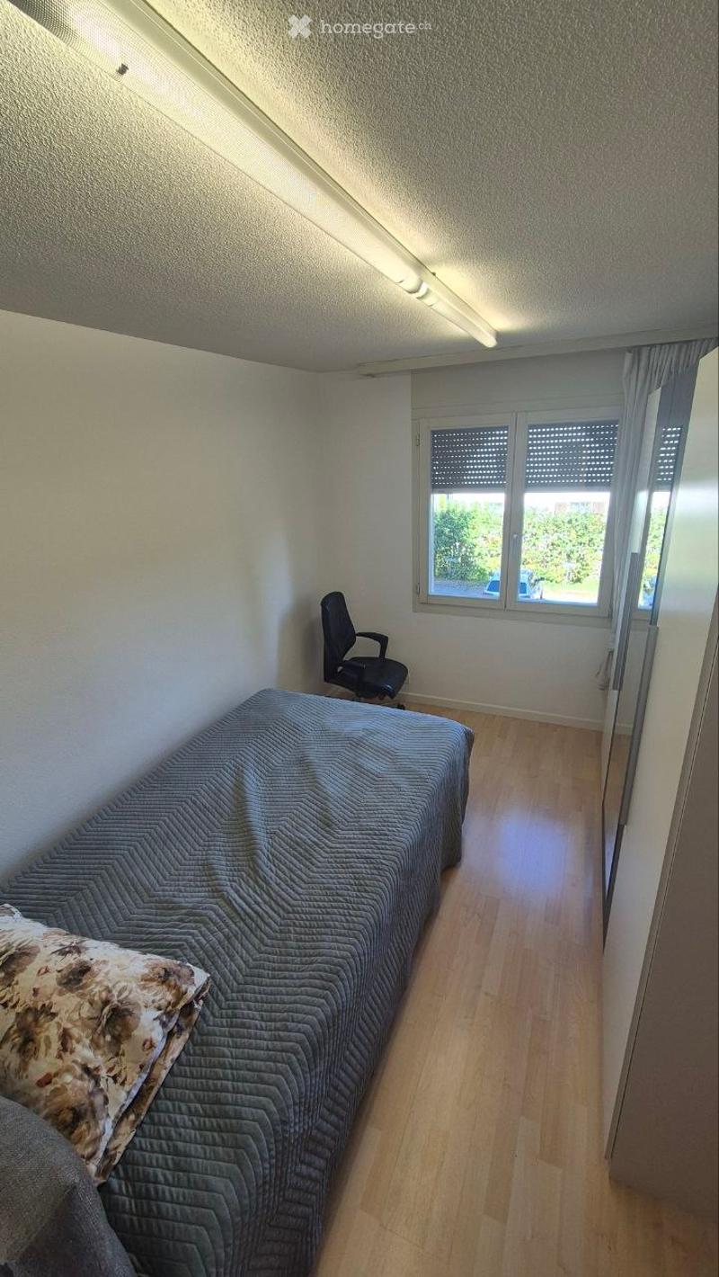 A small, well-lit bedroom with a grey quilted bed, a decorative pillow, a black office chair, and wooden flooring.
