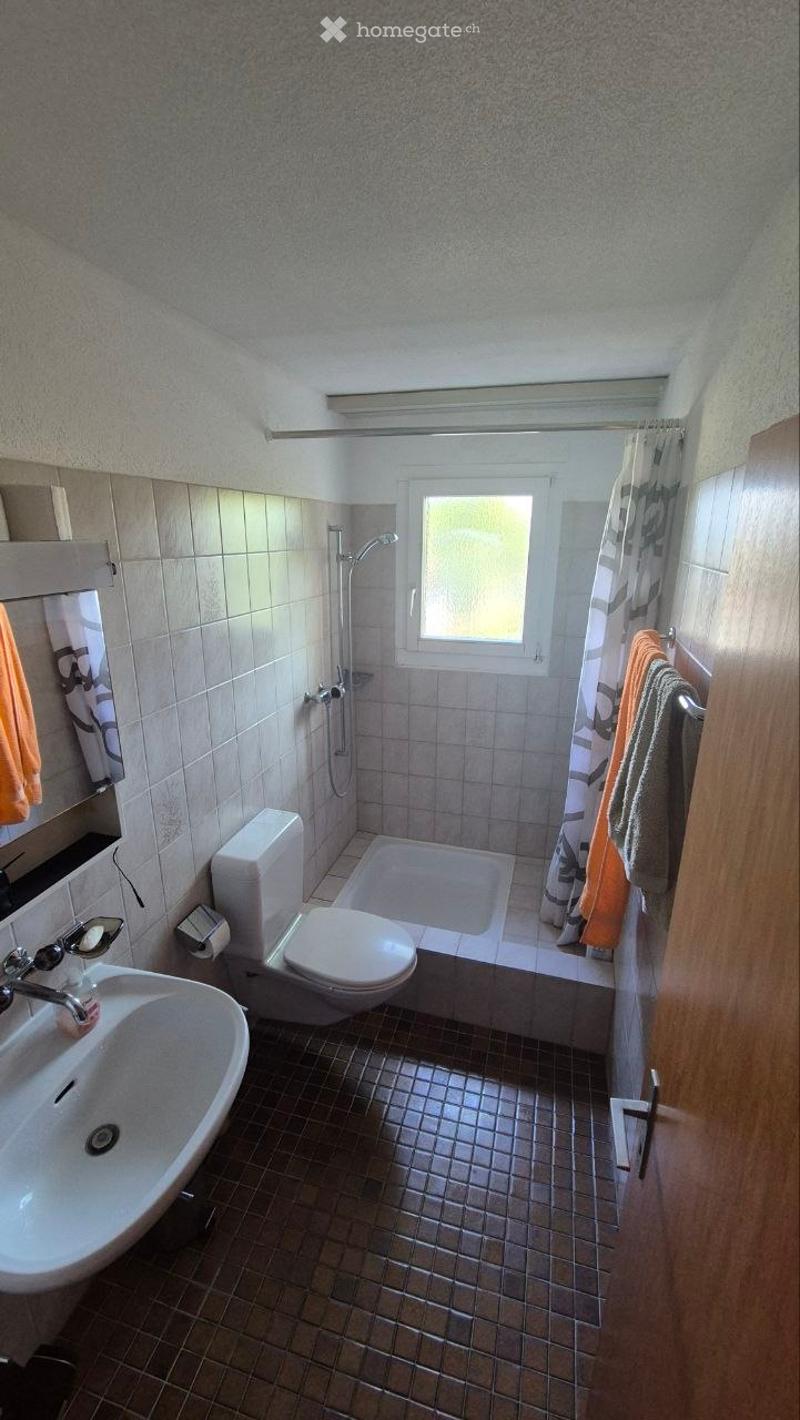 Bathroom with a sink, toilet, bathtub, shower, and towels hanging on the wall. The room has tiled walls and a window letting in natural light.
