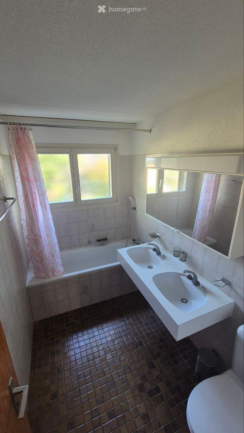 Bathroom with a bathtub, double sink, mirror, and toilet. Features tiled walls and floor, a window with a shower curtain, and a towel rack.