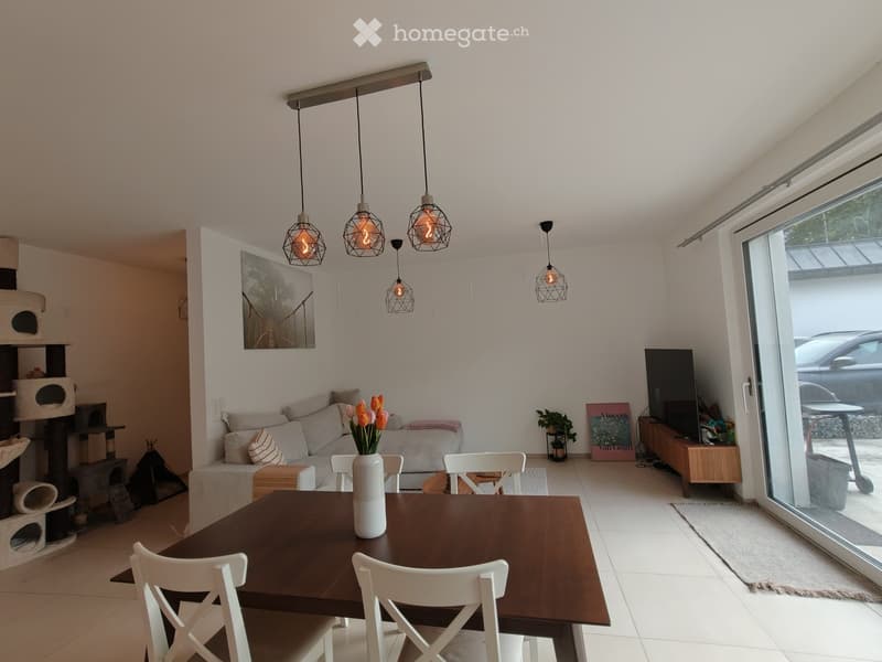 Bright living room with white walls, modern pendant lights, a sofa, a dining table with chairs, a cat tree, a TV, and a vase with flowers on the table.