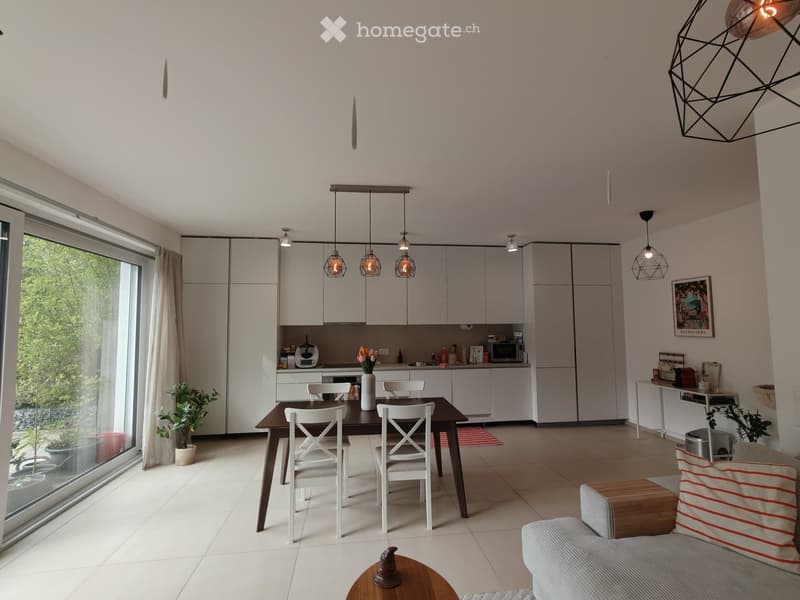 Modern open-plan living and dining area with white cabinets, pendant lights, a sofa, coffee table, dining table with chairs, and a kitchen area with appliances.