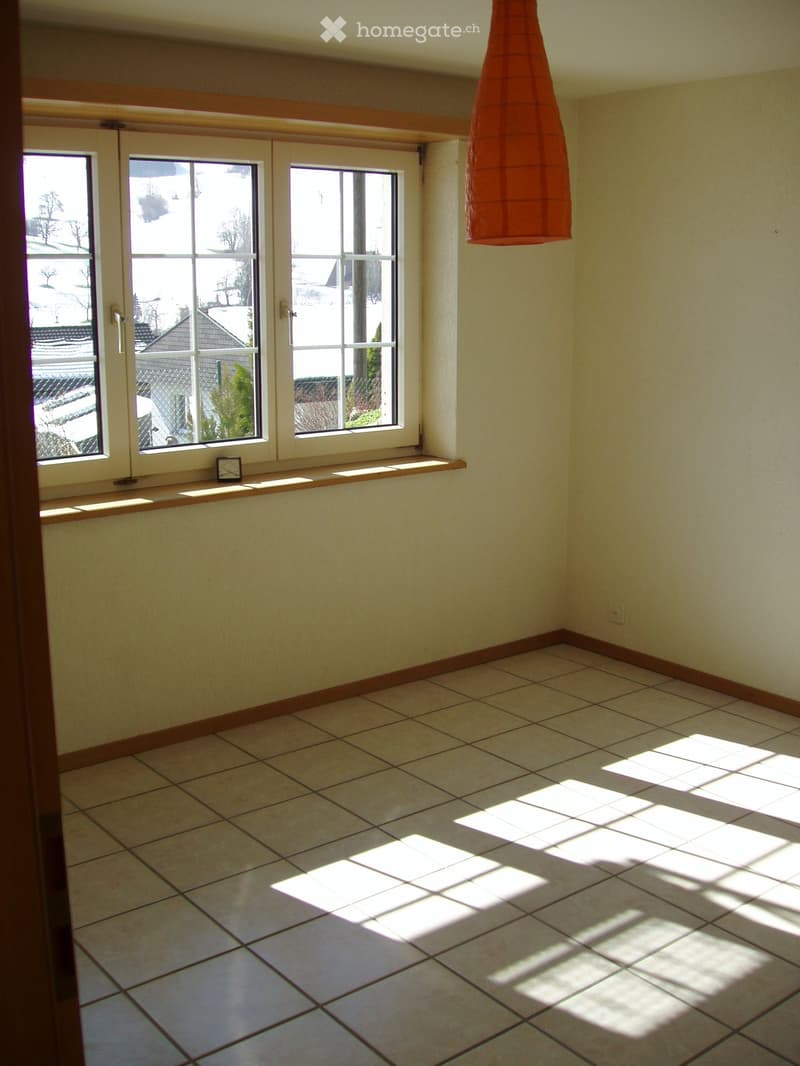 Empty room with tiled floor, large windows, and a small shelf with a clock. Natural light streams in, casting shadows on the floor.