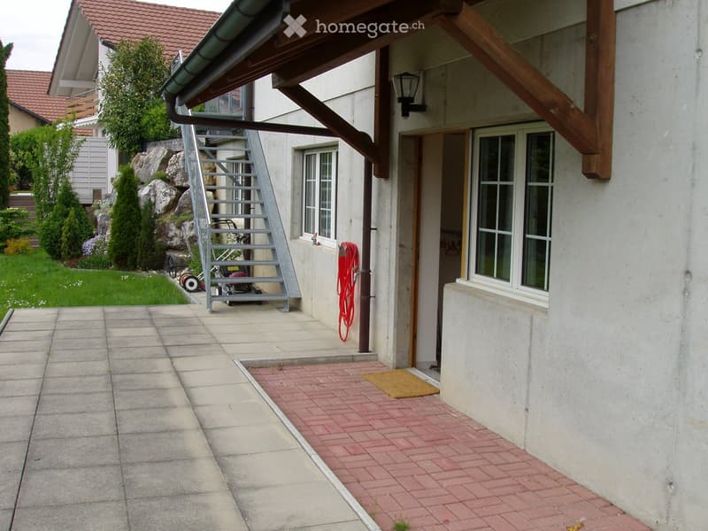 Exterior view of a two-story house with a red-tiled roof, a covered entrance area, and a small garden with rocks and plants.