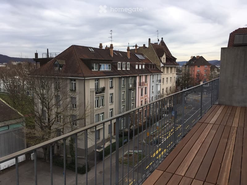 Balcony with wooden decking and metal railing overlooking a street lined with multi-story residential buildings, some with balconies and chimneys.