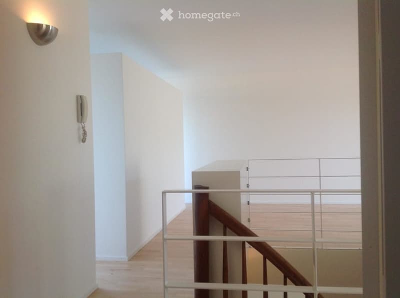 Bright hallway with wooden flooring, white walls, and a modern staircase with a metal railing. A wall-mounted light fixture and a telephone are visible on the left side.
