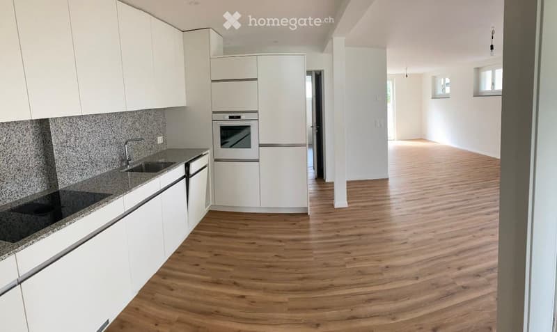 Empty room with wooden flooring, white walls, and a doorway leading to a modern kitchen area with white cabinets, granite countertops, and built-in appliances.