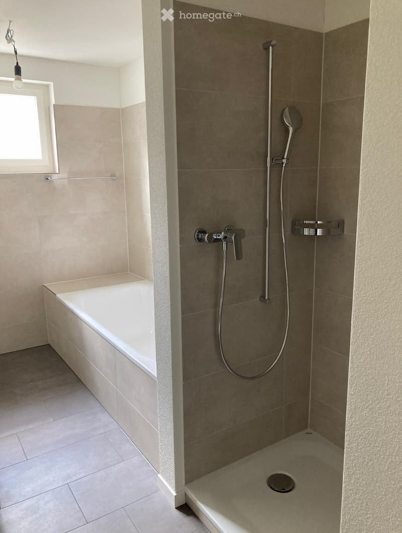 Modern bathroom with a bathtub and a separate shower area, featuring beige tiles and a window for natural light.