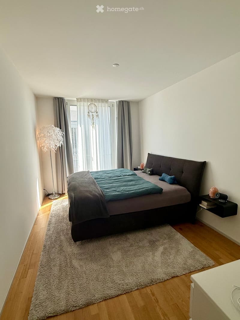 Modern bedroom with a large bed, nightstand, floor lamp, and a dreamcatcher hanging from the ceiling. The room features wooden flooring and a large window with curtains.